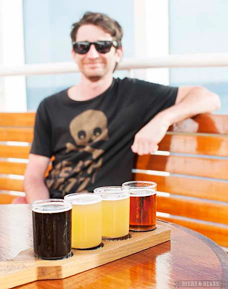 Four beer tastings sitting on a wooden holder with Randy in the background wearing sunglasses and a black shirt