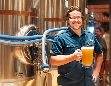 Brewmaster Colin Presby holding a pitcher of IPA in front of beer tanks on Carnival Vista